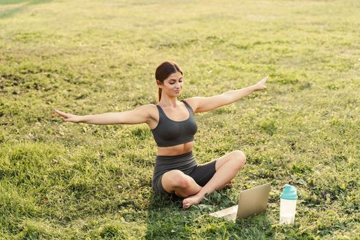 Yoga meditation online with laptop on the grass in green park. Fit girl training outdoor. Relaxation and meditating