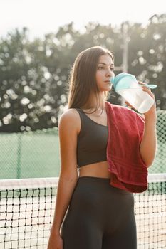 Fit sports model is resting after training and drinking water from a bottle. Sports lifestyle. Beautiful girl posing on the tennis court