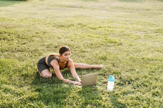 Fit girl with laptop on the green grass. Fitness trainer is chatting online with clients and explain exercises. Sport lifestyle