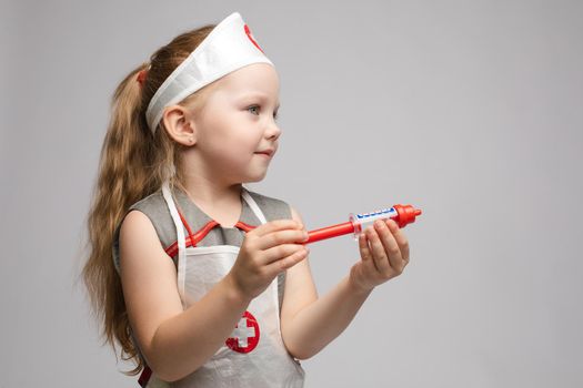 Little cute funny girl playing wearing doctor uniform holding toy syringe looking at camera. Smiling baby female child in glasses posing in nurse garb at white studio background medium long shot