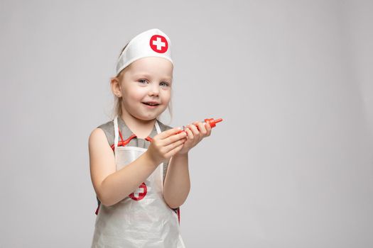 Little cute funny girl playing wearing doctor uniform holding toy syringe looking at camera. Smiling baby female child in glasses posing in nurse garb at white studio background medium long shot