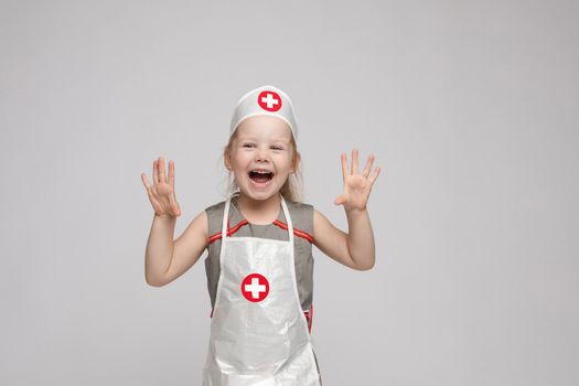 Stock photo of lovely little girl in white apron and a hat playing in a doctor. She is pointing at her hat with white cross in red circle. She is a doctor.