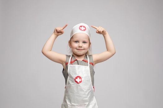 Stock photo of lovely little girl in white apron and a hat playing in a doctor. She is pointing at her hat with white cross in red circle. She is a doctor.