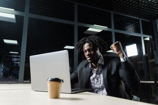 Happy black man with clenched fists is looking on the screen of laptop and rejoice finishing his job