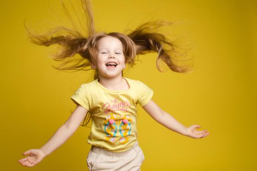 Front view of beautiful longhaired girl in cute shirt playing with hair and twirling in studio. Sunny little child looking at camera and posing on yellow isolated background. Concept of childhood.