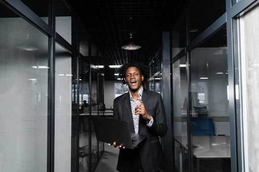 Cheerful black business manager with laptop is pointing fingers up and smiling in office