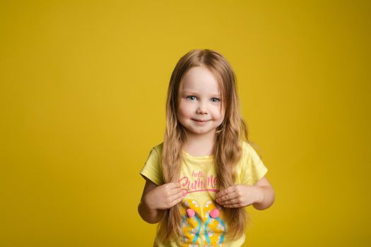 Stock photo portrait of cute and sweet little girl with long fair hair in yellow t-shirt with print holding her hair in hands and smiling at camera. She is isolated on yellow background. Smiling at camera shyly.