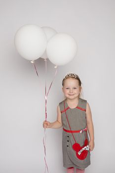 Stock photo of pretty little girl in nice dress and princess crown holding white air balloons and smiling at camera. Studio shot. Isolate on grey background.