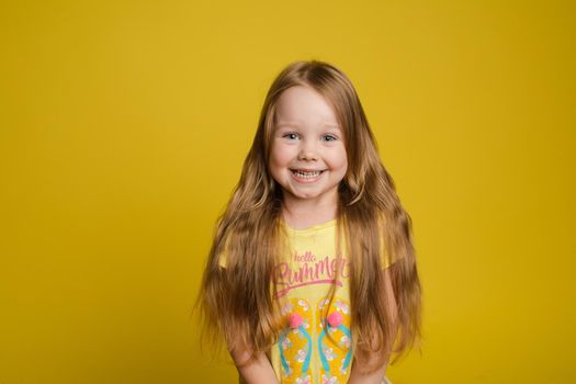 Portrait of beautiful little girl with long hair smiling posing isolated at yellow studio background medium shot. Happy cute European female child having fun looking at camera feeling positive emotion