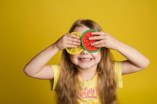 Front view of happy girl standing on yellow isolated background and closing eyes with toys. Cheerful longhaired child laughing and playing hide and seek. Concept of game and happiness.