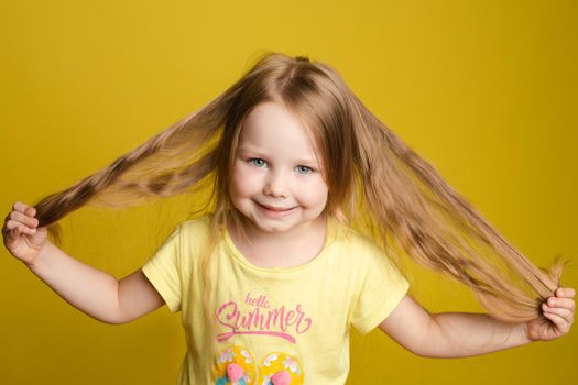 Front view of beautiful longhaired girl in cute shirt playing with hair and twirling in studio. Sunny little child looking at camera and posing on yellow isolated background. Concept of childhood.