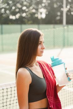 Fit sports model is resting after training and drinking water from a bottle. Sports lifestyle. Beautiful girl posing on the tennis court