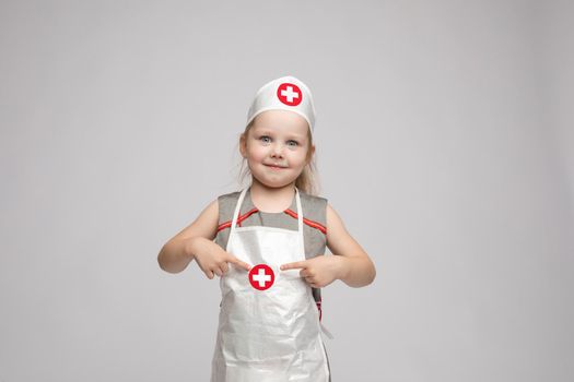 Stock photo of lovely little girl in white apron and a hat playing in a doctor. She is pointing at her hat with white cross in red circle. She is a doctor.