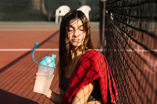 Athletic fit girl drinks water from a bottle. Rest after training on the tennis court