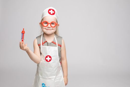 Little cute funny girl playing wearing doctor uniform holding toy syringe looking at camera. Smiling baby female child in glasses posing in nurse garb at white studio background medium long shot