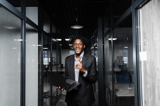 Cheerful black hipster man with laptop is pointing fingers up and smiling in office