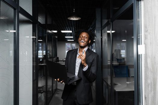 Cheerful black business manager with laptop is pointing fingers up and smiling in office