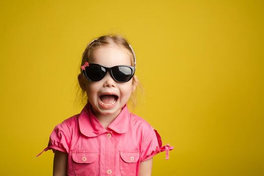 Front view of funny girl in pink shirt wearing glasses and shouting. Pretty little child looking at camera and posing on yellow isolated background. Concept of summer outfit and childhood.