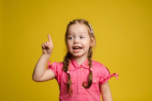 Stock photo of cute little girl with hair accessories wearing sunglasses and pink dress showing the direction with her index finger. Isolate on yellow background.