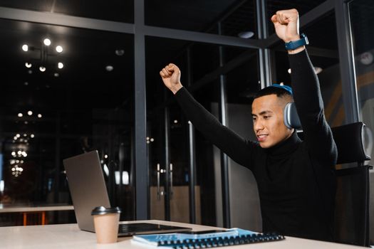 Asian man raises his hands and win bet online and earn money. Happy filipino guy with laptop and headphones celebrating victory in game.