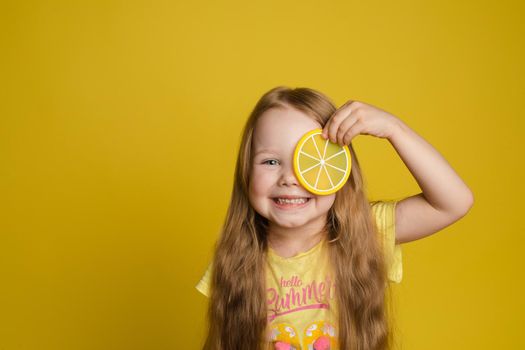 Portrait of smiling cute little girl holding slice of lemon near eye looking at camera medium close-up. Happy female beautiful child having fun posing isolated at yellow studio background