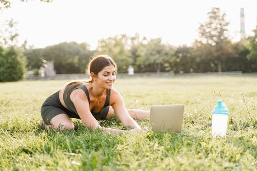 Fit girl with laptop on the green grass. Fitness trainer is chatting online with clients and explain exercises. Sport lifestyle