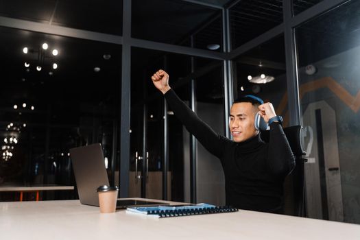 Asian man raises his hands and win bet online and earn money. Happy filipino guy with laptop and headphones celebrating victory in game.