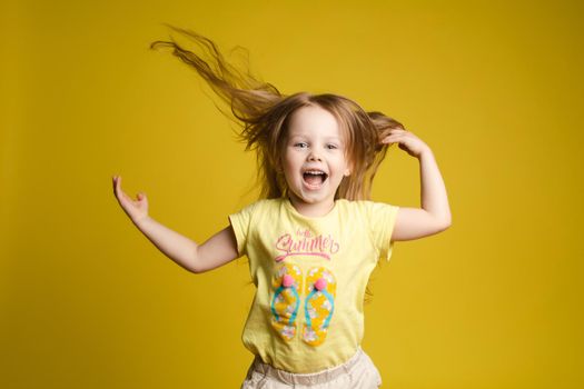 Front view of beautiful longhaired girl in cute shirt playing with hair and twirling in studio. Sunny little child looking at camera and posing on yellow isolated background. Concept of childhood.