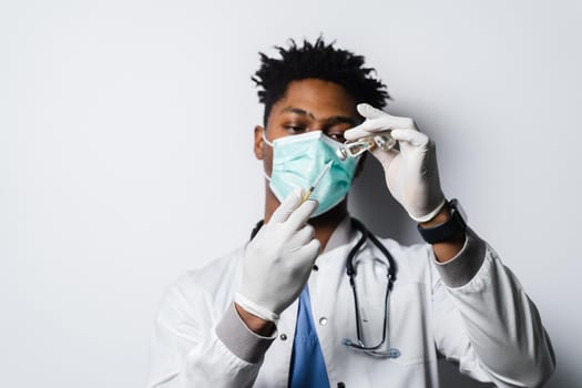 African doctor in a medical mask prepares to give an injection of a coronavirus covid-19 vaccine. Black man in medical mask is making vaccination on white background