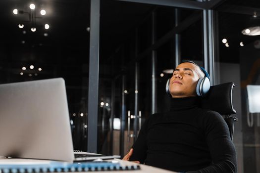 Overtime working. Asian man with closed eyes is resting at work at laptop. Tired male programmer taking a break due to overtime work.