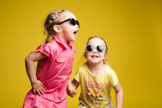 Two happy beautiful stylish little girl wearing sunglasses posing isolated at yellow studio background medium shot. Adorable laughing female child friend enjoying friendship having positive emotion