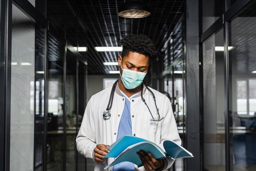 Black medical student reads books and prepares for an exam. African in a medical robe and mask looks at the notes