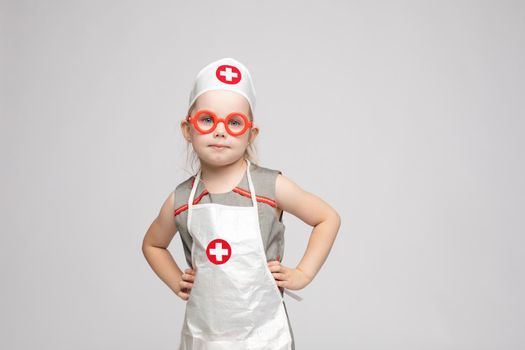 Stock photo of lovely little girl in white apron and a hat playing in a doctor. She is pointing at her hat with white cross in red circle. She is a doctor.