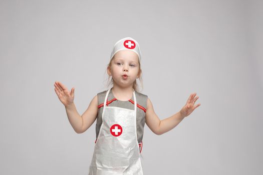 Stock photo of lovely little girl in white apron and a hat playing in a doctor. She is pointing at her hat with white cross in red circle. She is a doctor.