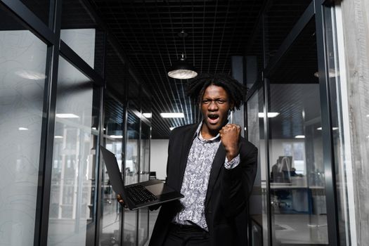 Happy black man clenched fists and rejoice finishing his online job on laptop. Working online in coworking space