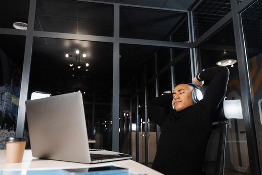 Asian man with closed eyes listening to relaxing music at work at laptop. Break at workplace for rest from overtime work