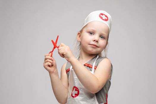 Stock photo portrait of cute little girl in doctor s hat and apron playing with plastic scissors. She is playing in doctor wearing medical uniform. Smiling at camera.