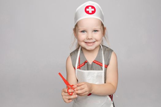 Stock photo portrait of cute little girl in doctor s hat and apron playing with plastic scissors. She is playing in doctor wearing medical uniform. Smiling at camera.