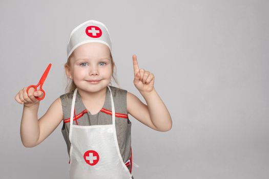 Stock photo portrait of cute little girl in doctor s hat and apron playing with plastic scissors. She is playing in doctor wearing medical uniform. Smiling at camera.