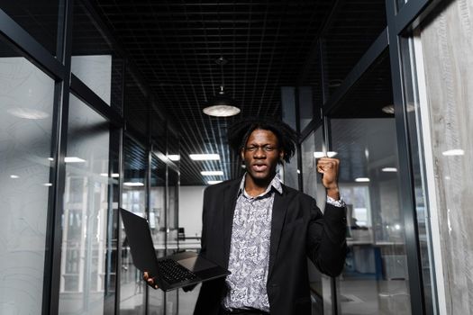 Happy black man clenched fists and rejoice finishing his online job on laptop. Working online in coworking space