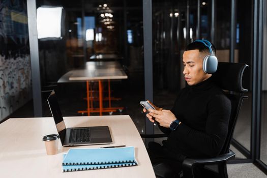 Asian man in headphones working online, listen to music and check his phone in office. Asian handsome guy with laptop in coworking space