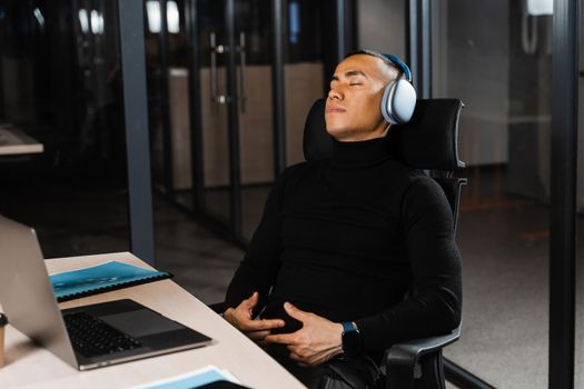 Asian man with closed eyes listening to relaxing music at work at laptop. Break at workplace for rest from overtime work