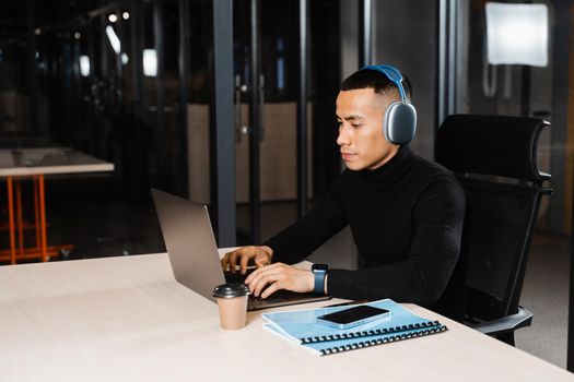 Asian man in headphones working online and listen to music in office. Asian handsome guy with laptop in coworking space