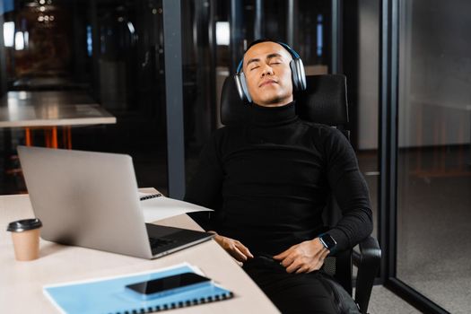 Overtime working. Asian man with closed eyes is resting at work at laptop. Tired male programmer taking a break due to overtime work.