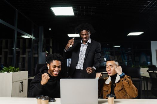 Multiethnic mixed race happy colleagues witl laptop. Black african and asian men raise their hands up, rejoice in victory and shout because they won an online bet