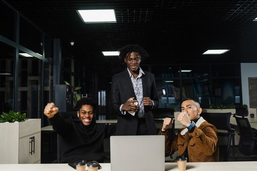 Emotional and joyful black Africans and Asians teamwork together and rejoice at end of online project. Multiethnic colleagues working on laptop, raising their hands, fists and shouting loudly