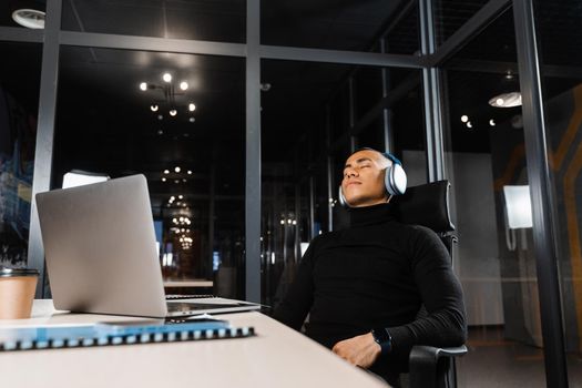 Asian man with closed eyes listening to relaxing music at work at laptop. Break at workplace for rest from overtime work