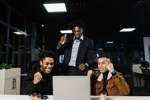 Multiethnic mixed race happy colleagues witl laptop. Black african and asian men raise their hands up, rejoice in victory and shout because they won an online bet