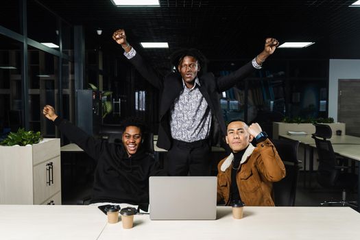 Happy and joyful multiethnic men raise their hands and fists and rejoice in winning in online casino on laptop. Teambuilding of black Africans and Asian programmers