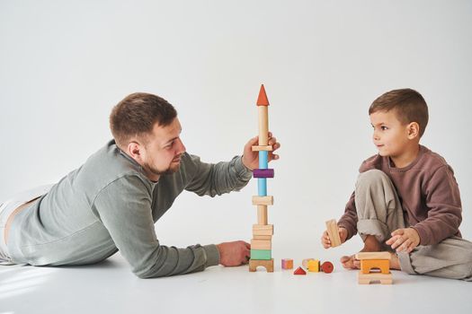 Caring dad helps his son to play on the floor on white background. Father and child build tower of colorful wooden bricks and have fun together
